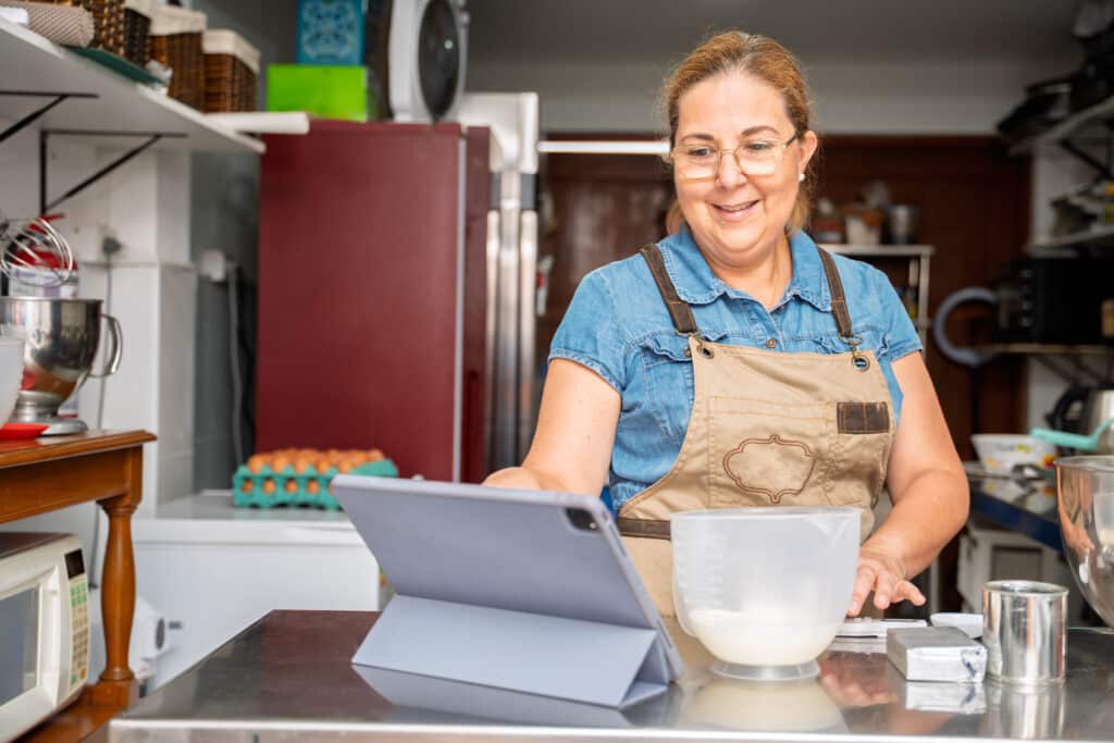 About 1 Stephanie the chef at My Dessert Spot smiling in her kitchen while looking at a tablet, preparing dessert ingredients.